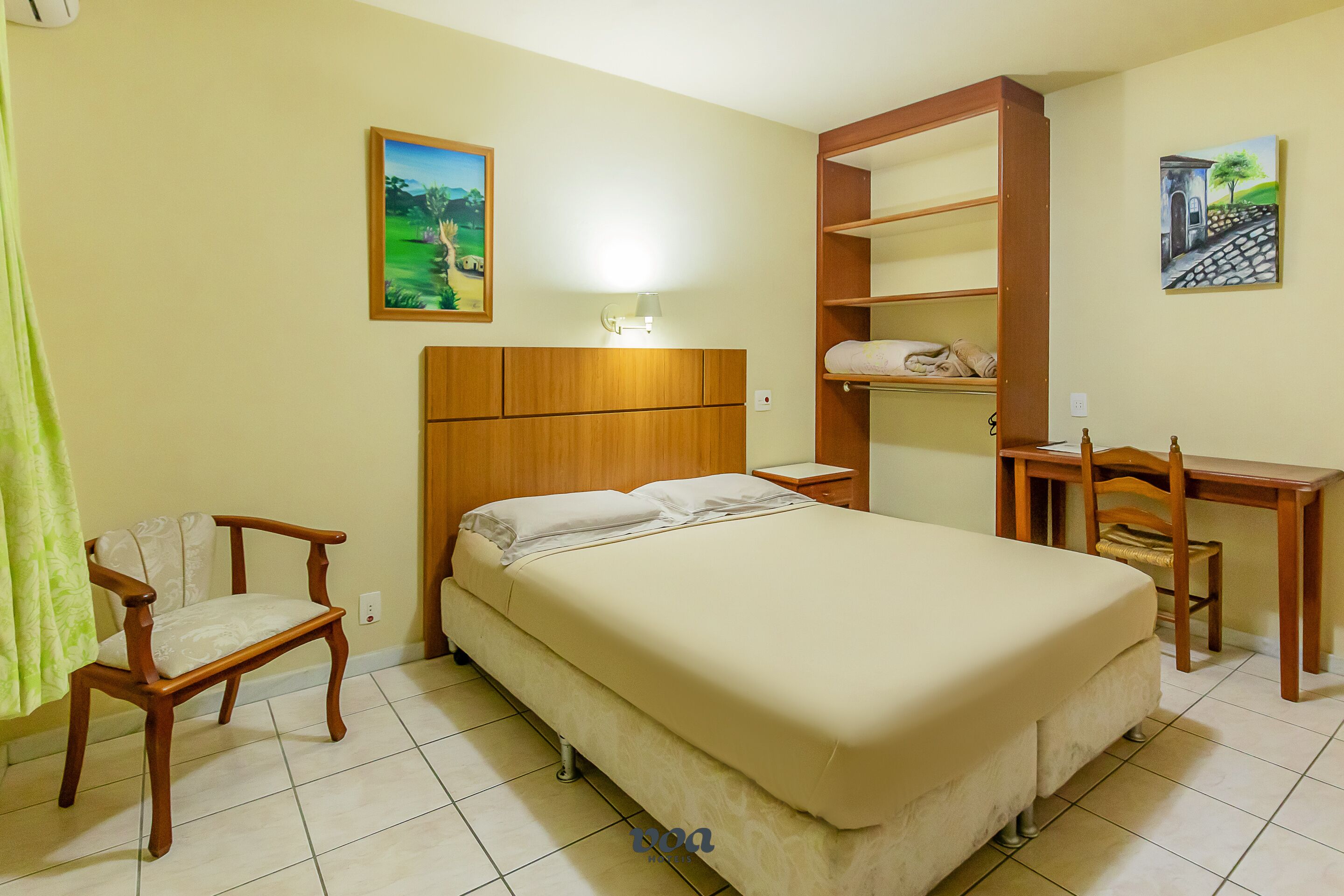 A hotel room with a bed, a wooden chair, a desk, and a shelf unit.