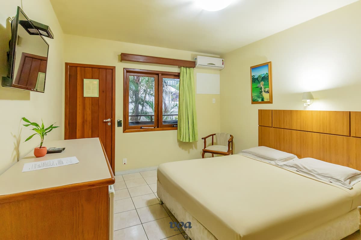A hotel room with a bed, a desk with a plant, a chair, a window with curtains, and an air conditioning unit.