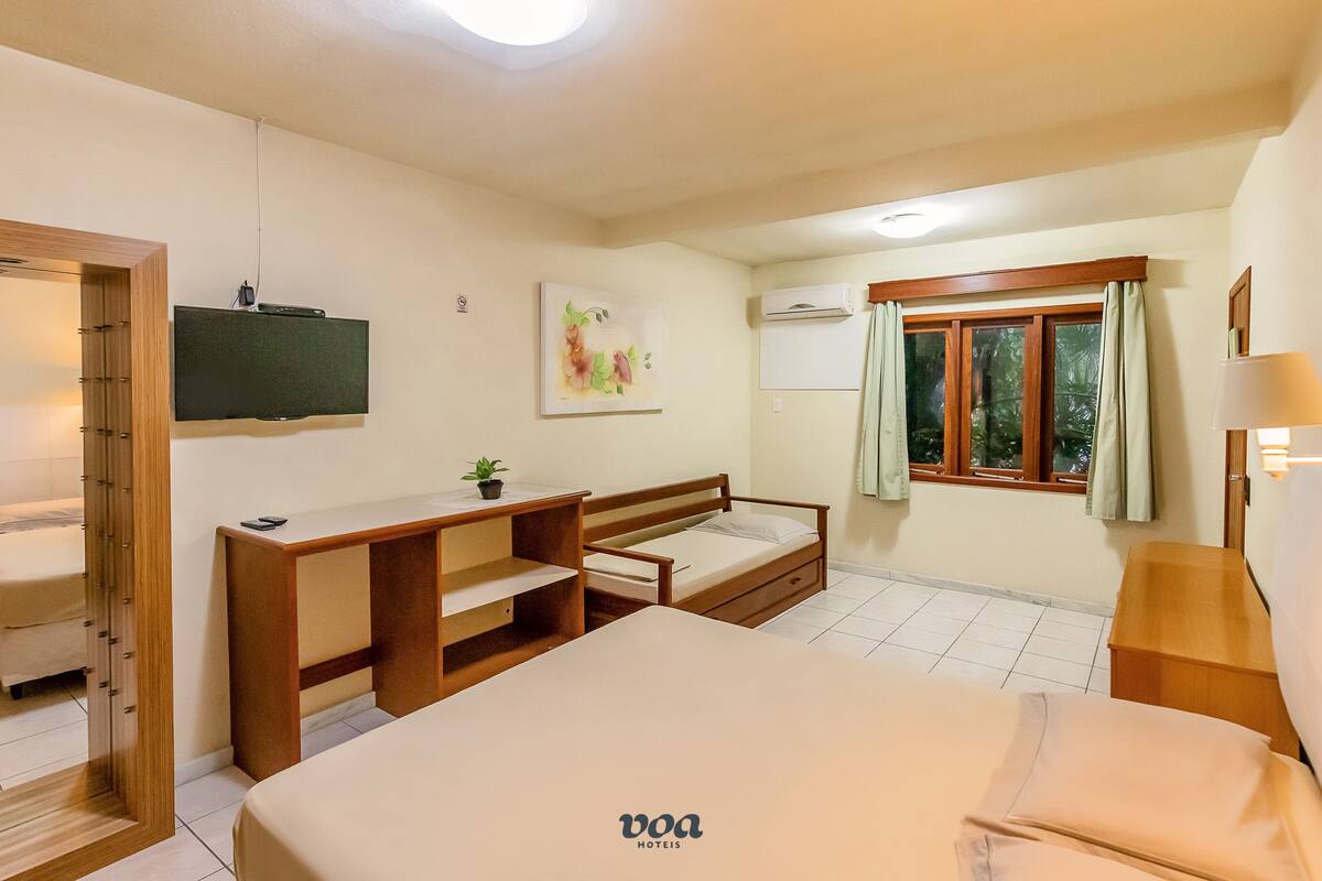 A hotel room with a bed, a desk, a chair, a television, a window with curtains, and a wall-mounted air conditioner.