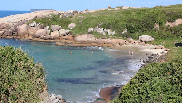 A coastal landscape with large boulders, clear water, and a grassy hillside. - VOA Samuka Hotel (Florianópolis)