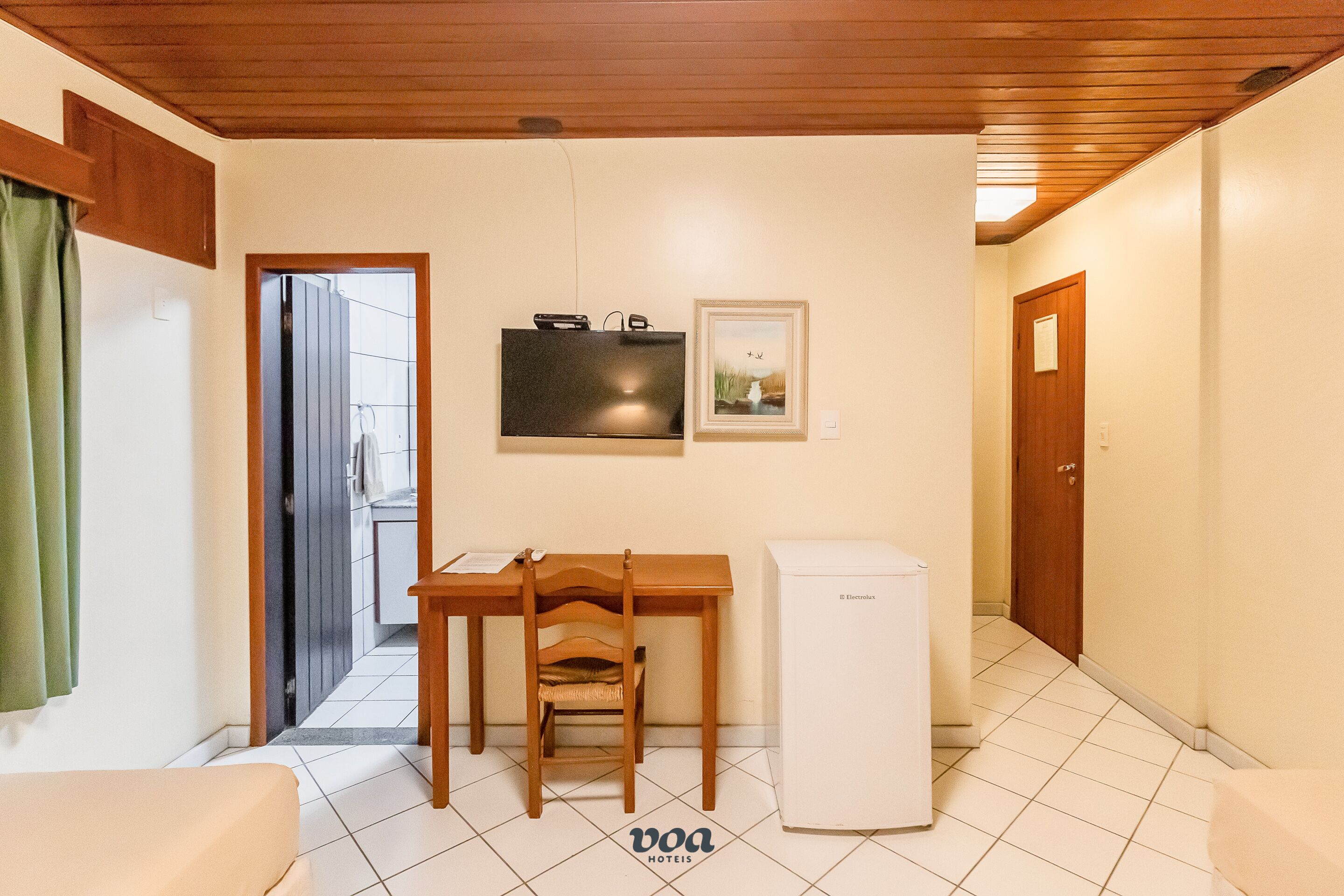 A compact room with a bed, a small table with a chair, a refrigerator, and a door leading to another room.