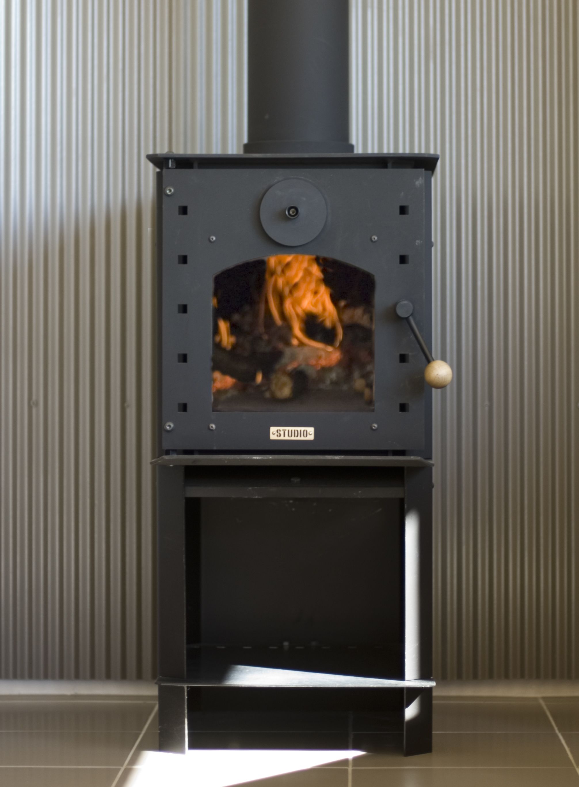 A black wood-burning stove with a visible fire inside, a control knob, and a label reading "STUDIO".