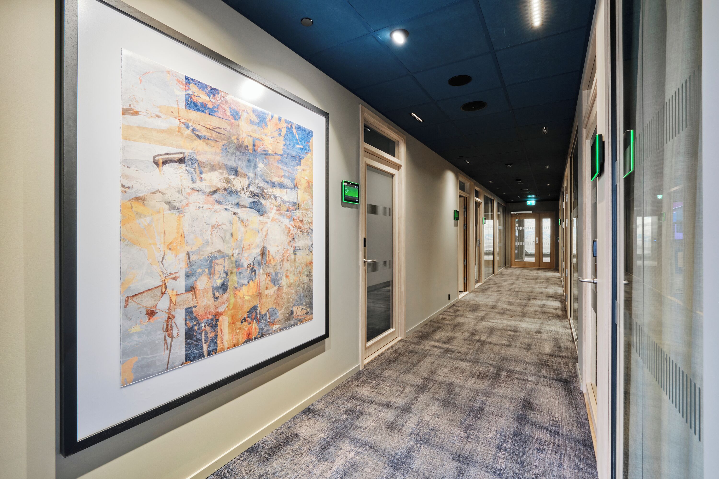A hallway with a large abstract painting, carpeted floors, glass walls, and doors with green signs.