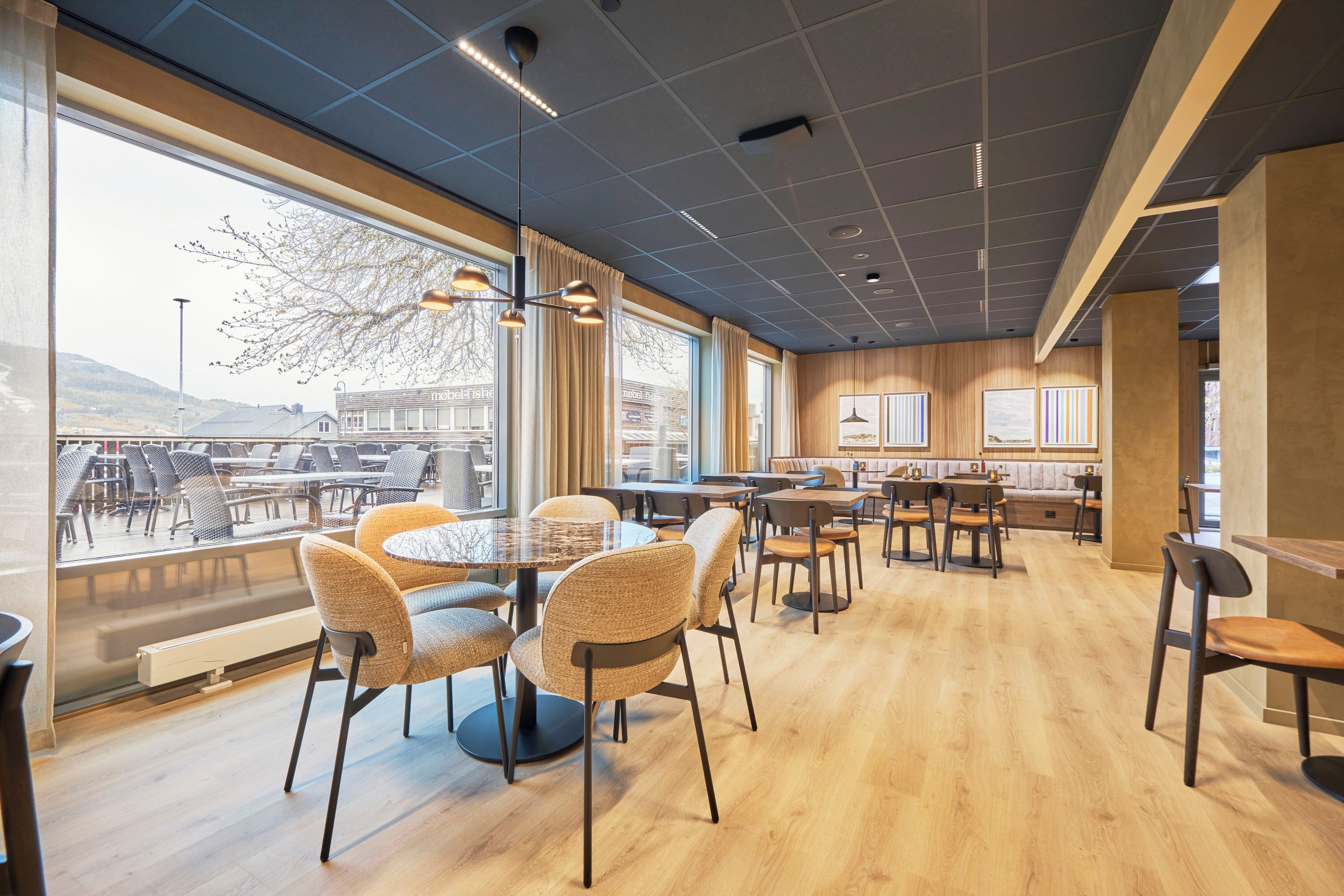 Et moderne spiseområde med runde borde og stole, store vinduer med udsigt og et træloft.