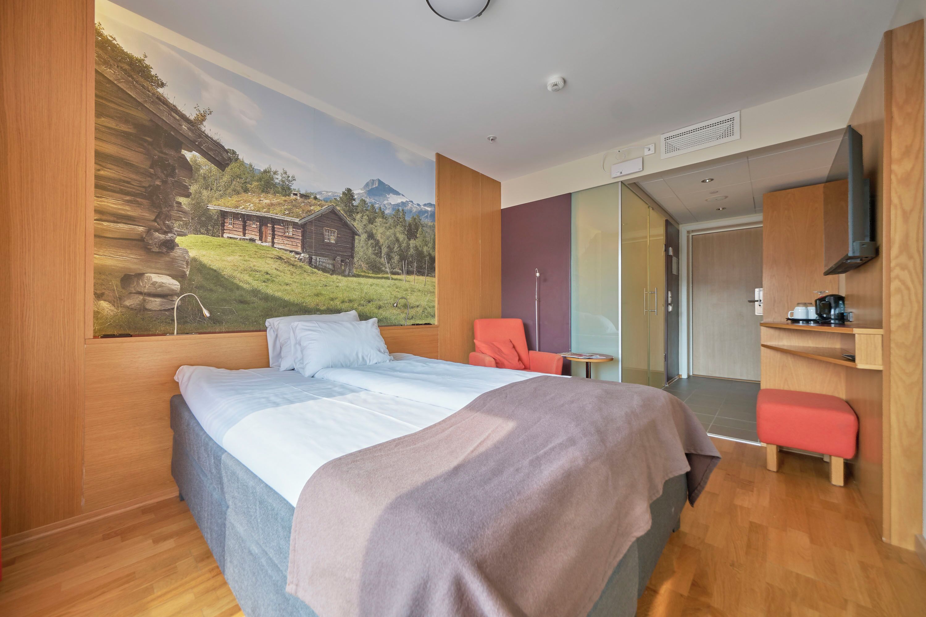 Una habitación de hotel con una cama, una silla, una mesita y un cuadro de un paisaje montañoso.