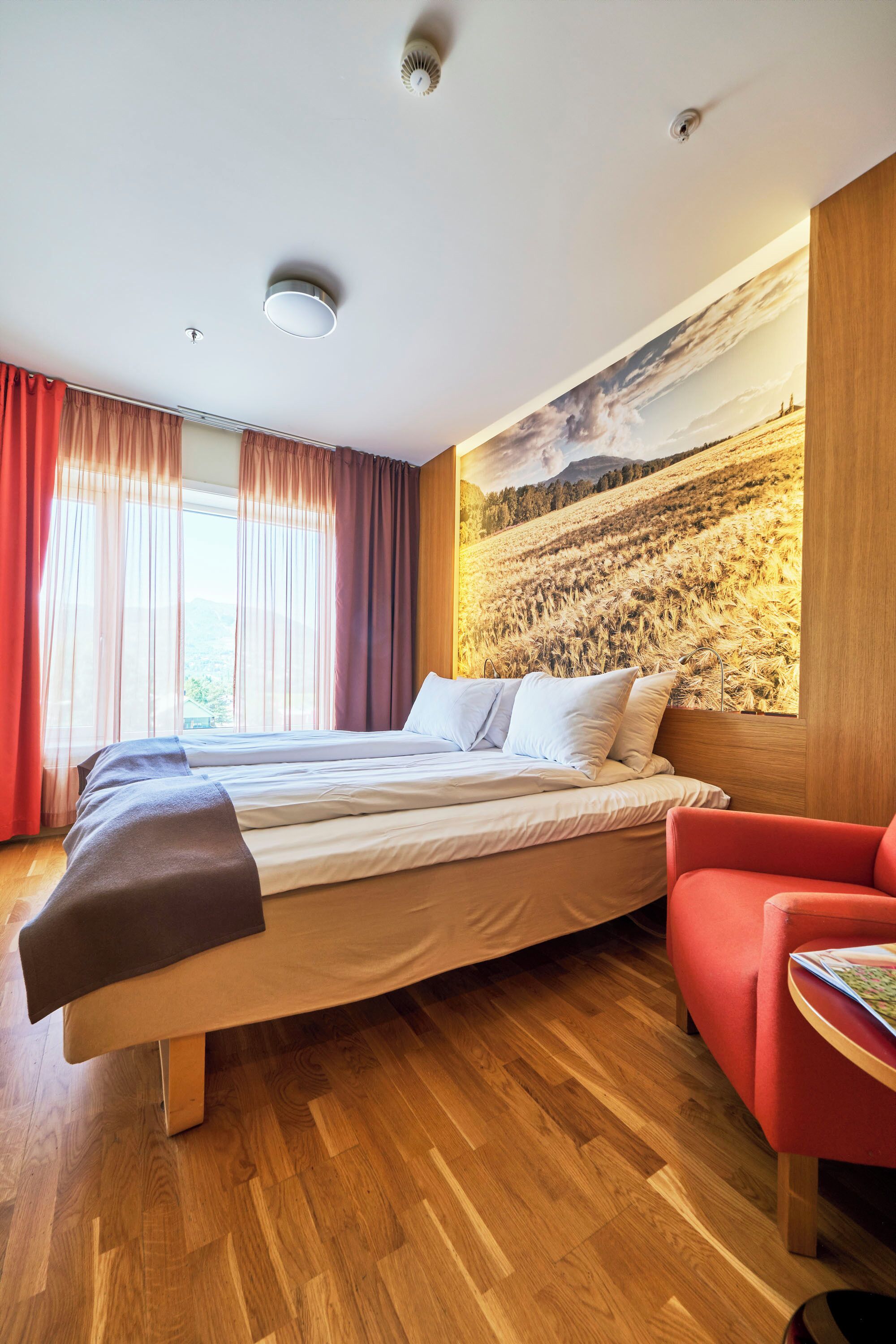 A bedroom with a bed, a red armchair, wooden flooring, and a large landscape painting on the wall.
