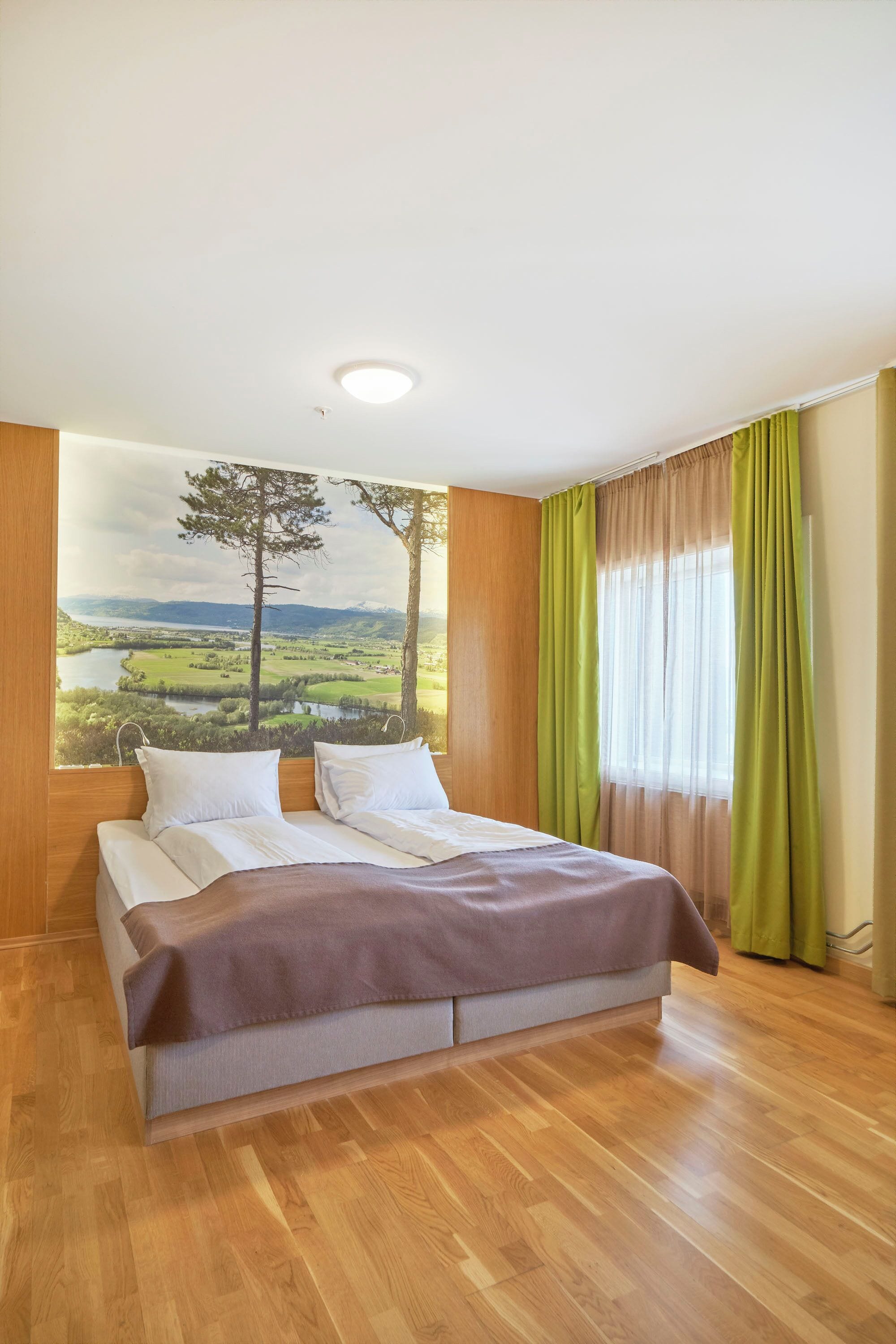 Une chambre à coucher avec un lit, une grande fenêtre avec des rideaux, un tableau représentant un paysage et un plancher en bois.