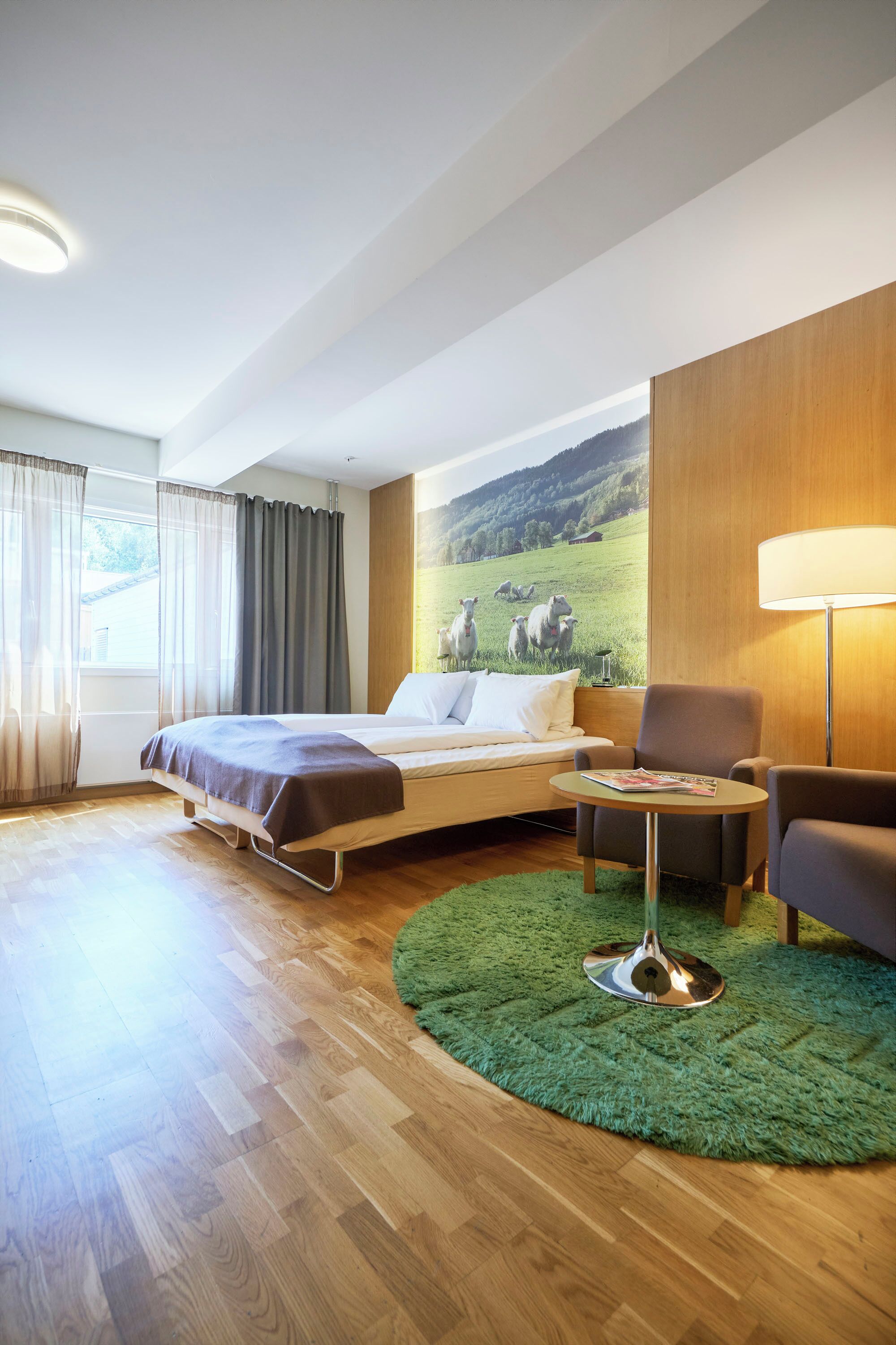 A hotel room with a bed, a sofa, a small table, a lamp, a green rug, and a large window with curtains.
