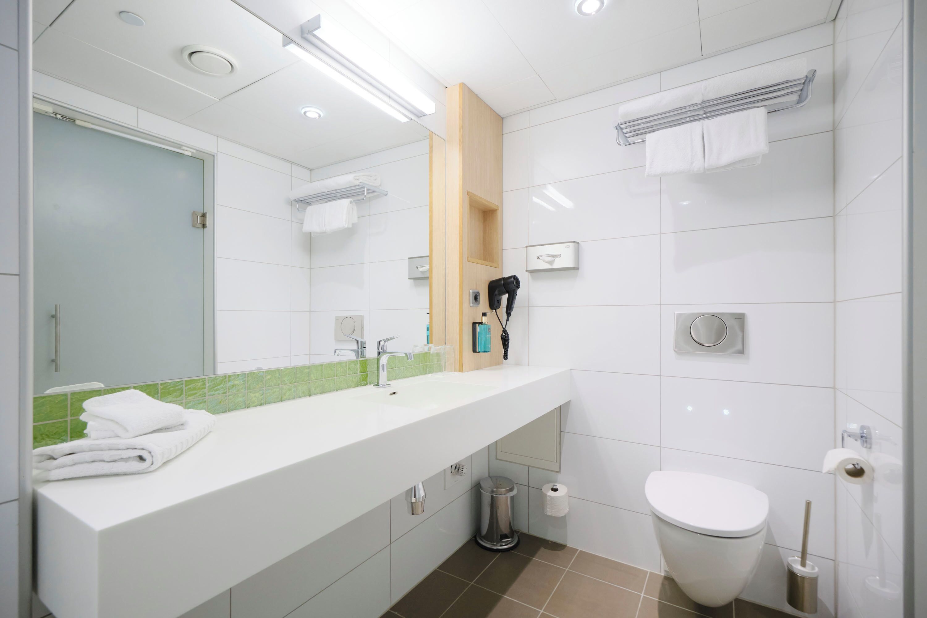A modern bathroom with a white sink, mirror, and toilet. There is a towel rack above the sink and a trash can next to the toilet.