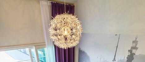 Een moderne woonkamer met een bank, stoelen, een bijzettafeltje, een hanglamp en een muurschildering van een stadsgezicht.