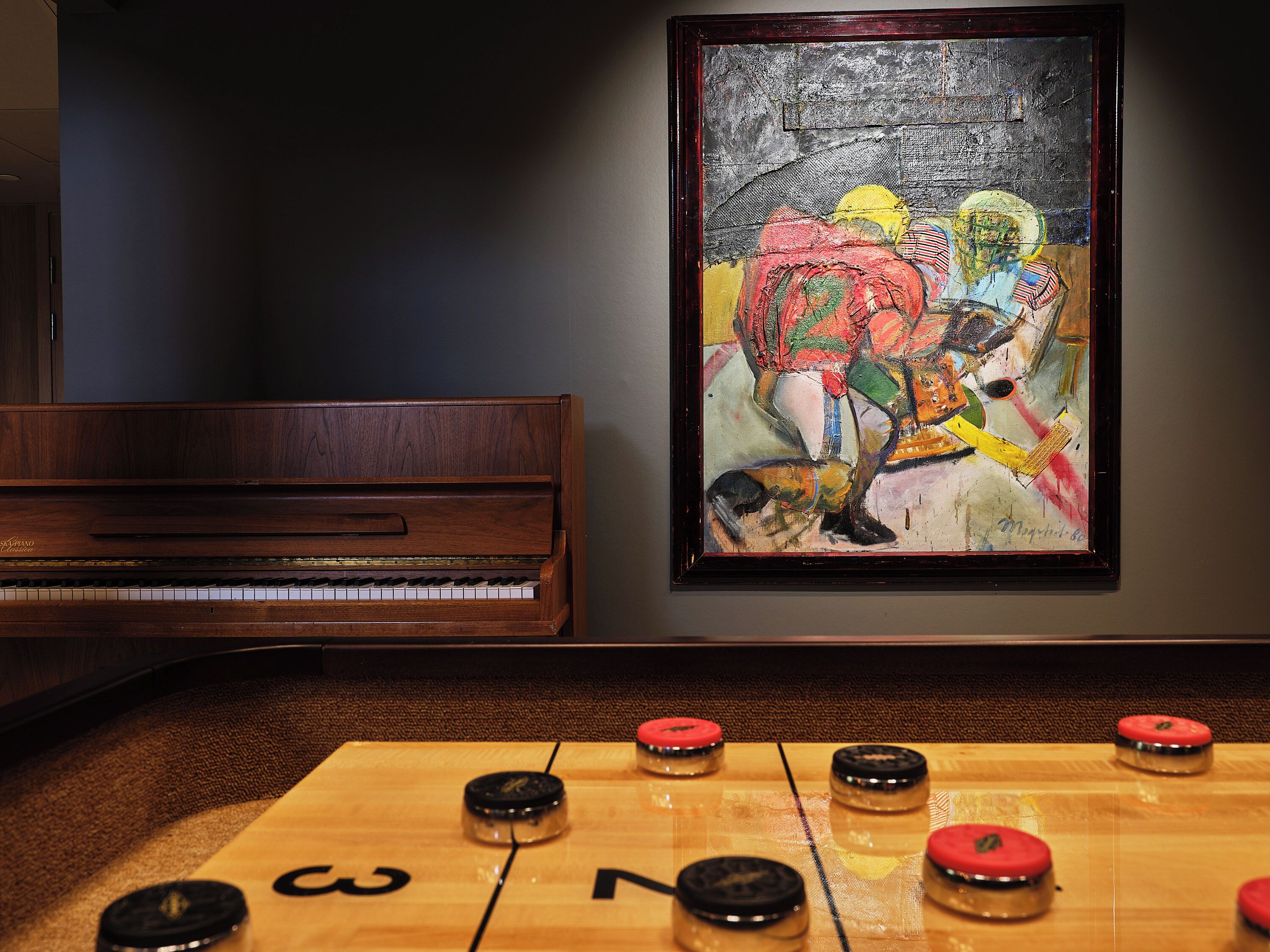 Una habitación con un piano, una mesa con discos de shuffleboard y un cuadro abstracto enmarcado en la pared.