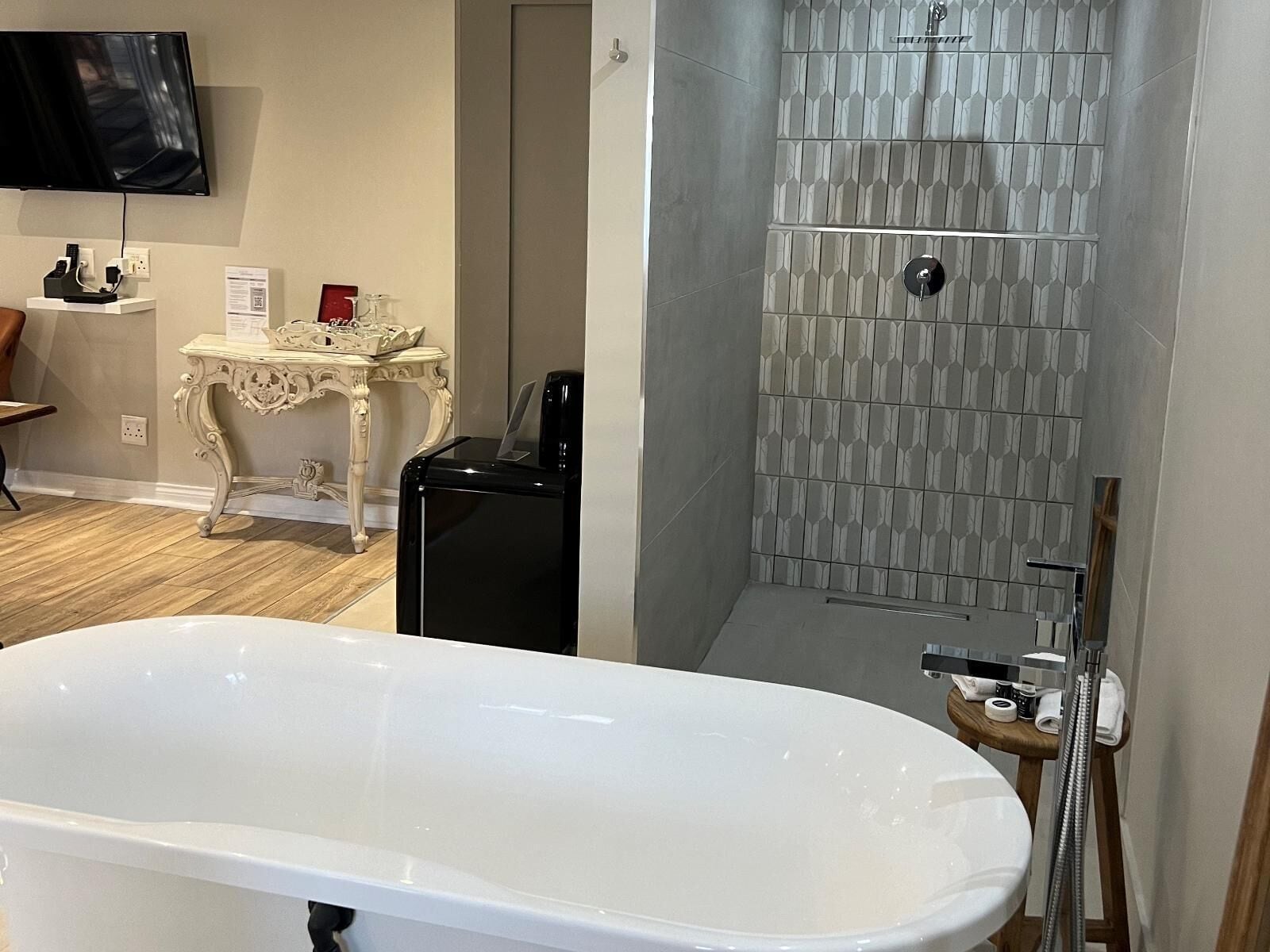 A bathroom with a freestanding bathtub, a shower with a patterned tile wall, a small table with a lamp, and a television mounted on the wall.