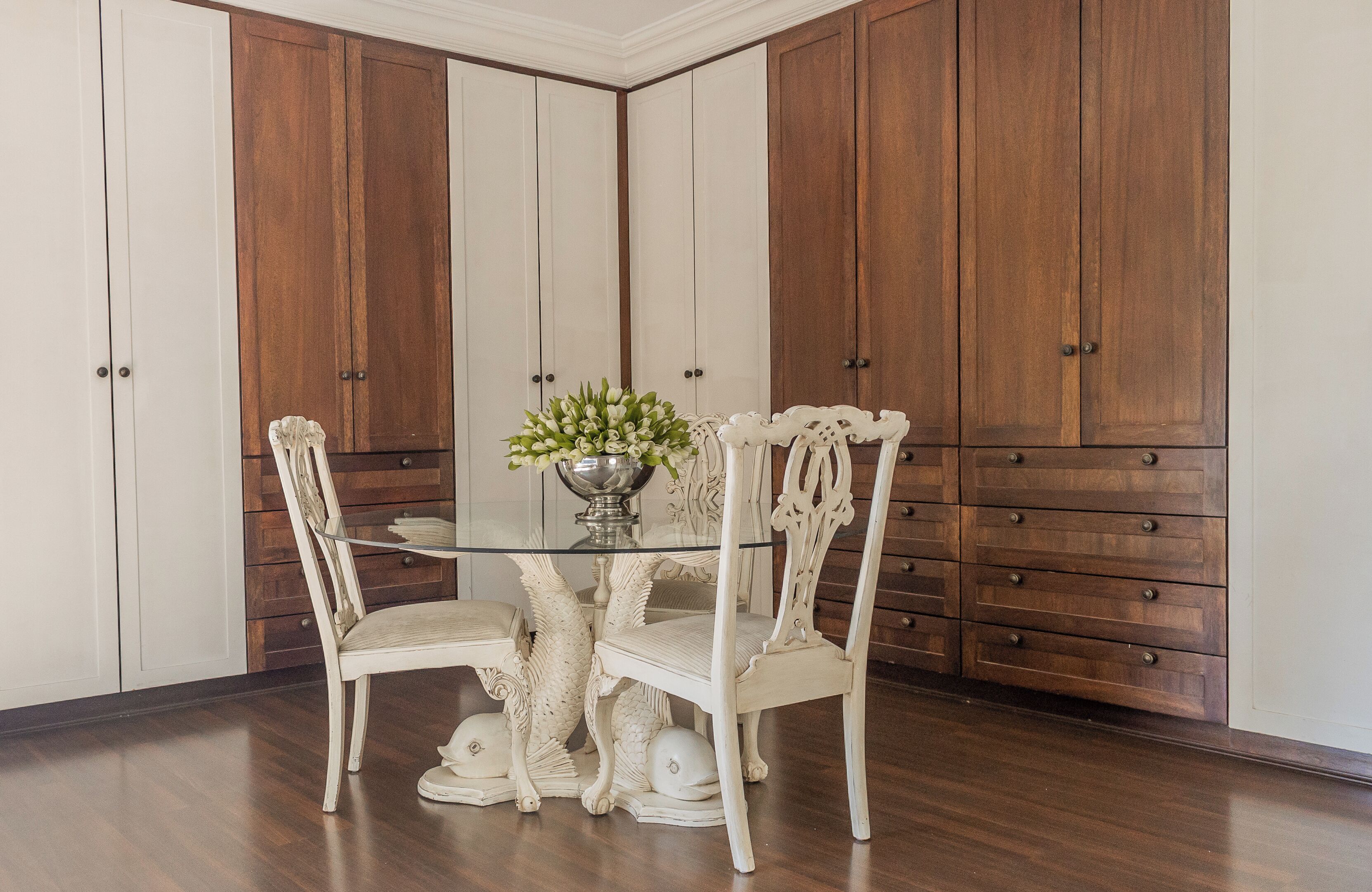 A dining area with a glass table surrounded by ornate chairs, a vase with flowers, and wooden cabinets.