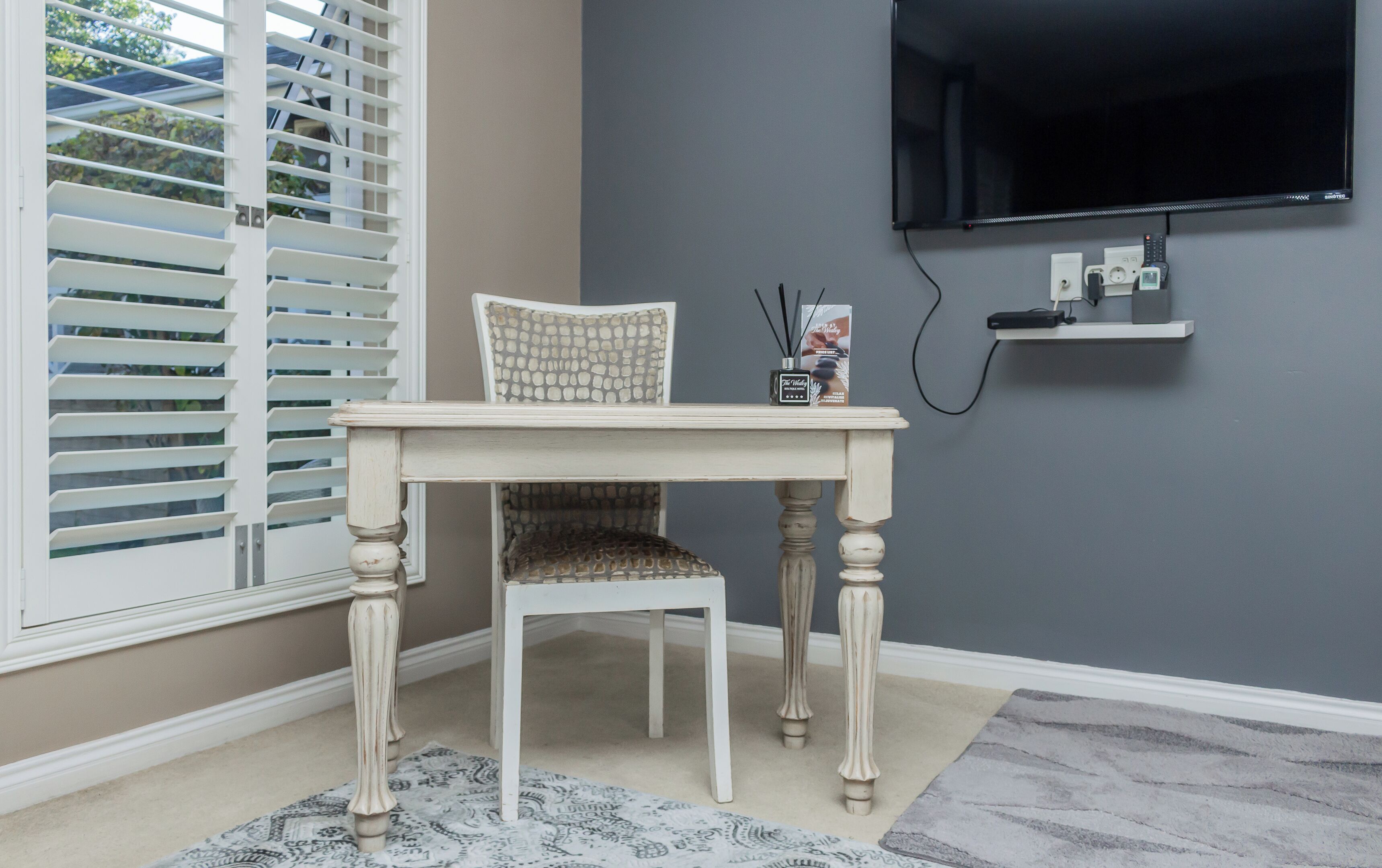 A room with a white table and chair, a grey wall, a mounted TV, and a small shelf with a remote control.