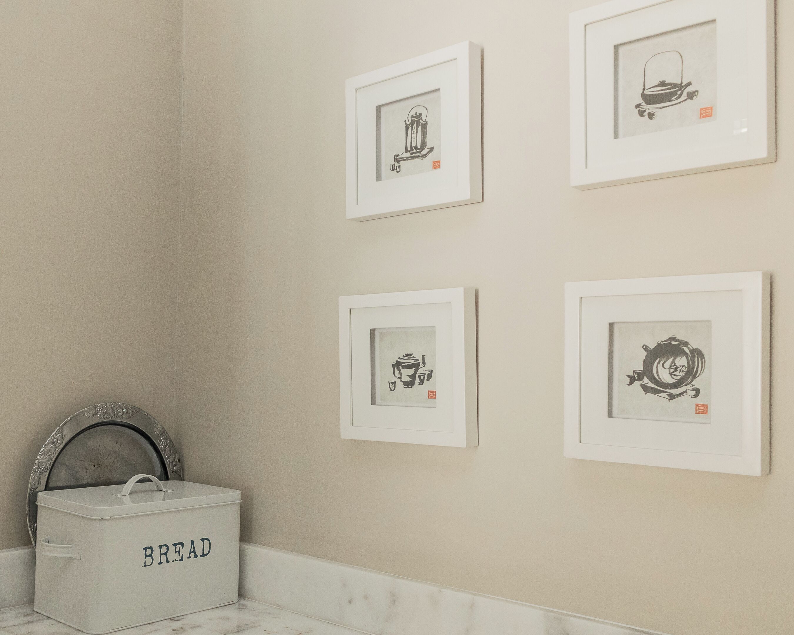 Une salle de bain avec quatre œuvres d’art encadrées accrochées au mur, une boîte à pain blanche portant l’inscription « PAIN » posée sur le sol, et un miroir décoratif doté d’un cadre orné.