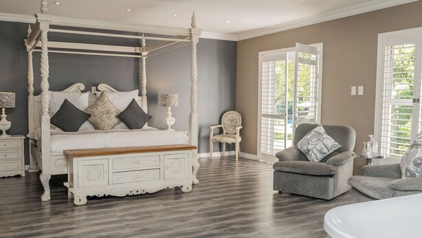 Un dormitorio con una cama con dosel, un banco de madera, un sillón gris y una mesita de noche blanca.