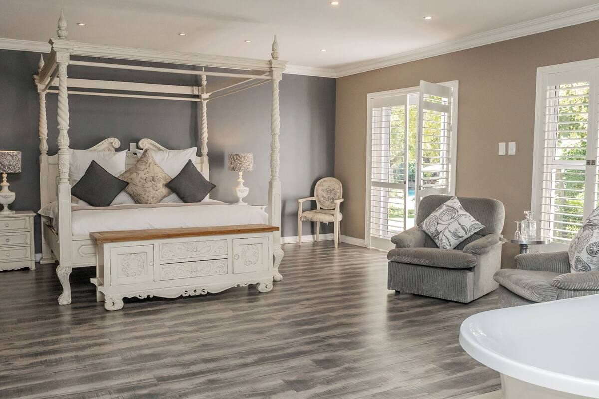 Une chambre à coucher avec un lit à baldaquin, un banc en bois, un fauteuil gris et une table de chevet blanche.