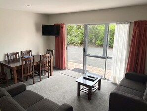 A modern living room with a dining area, a flat-screen TV mounted on the wall, and a sliding glass door leading to an outdoor area. - Jade Court Motor Lodge (Hokitika)