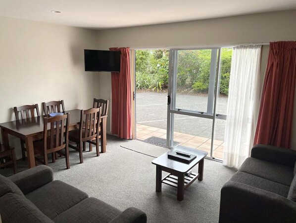 A modern living room with a dining area, a flat-screen TV mounted on the wall, and a sliding glass door leading to an outdoor area. - Jade Court Motor Lodge (Hokitika)