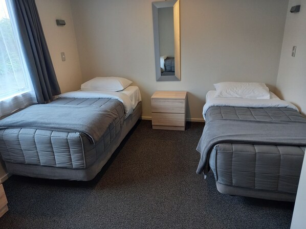 A hotel room with two beds, a small bedside table, a mirror, and a window with curtains.