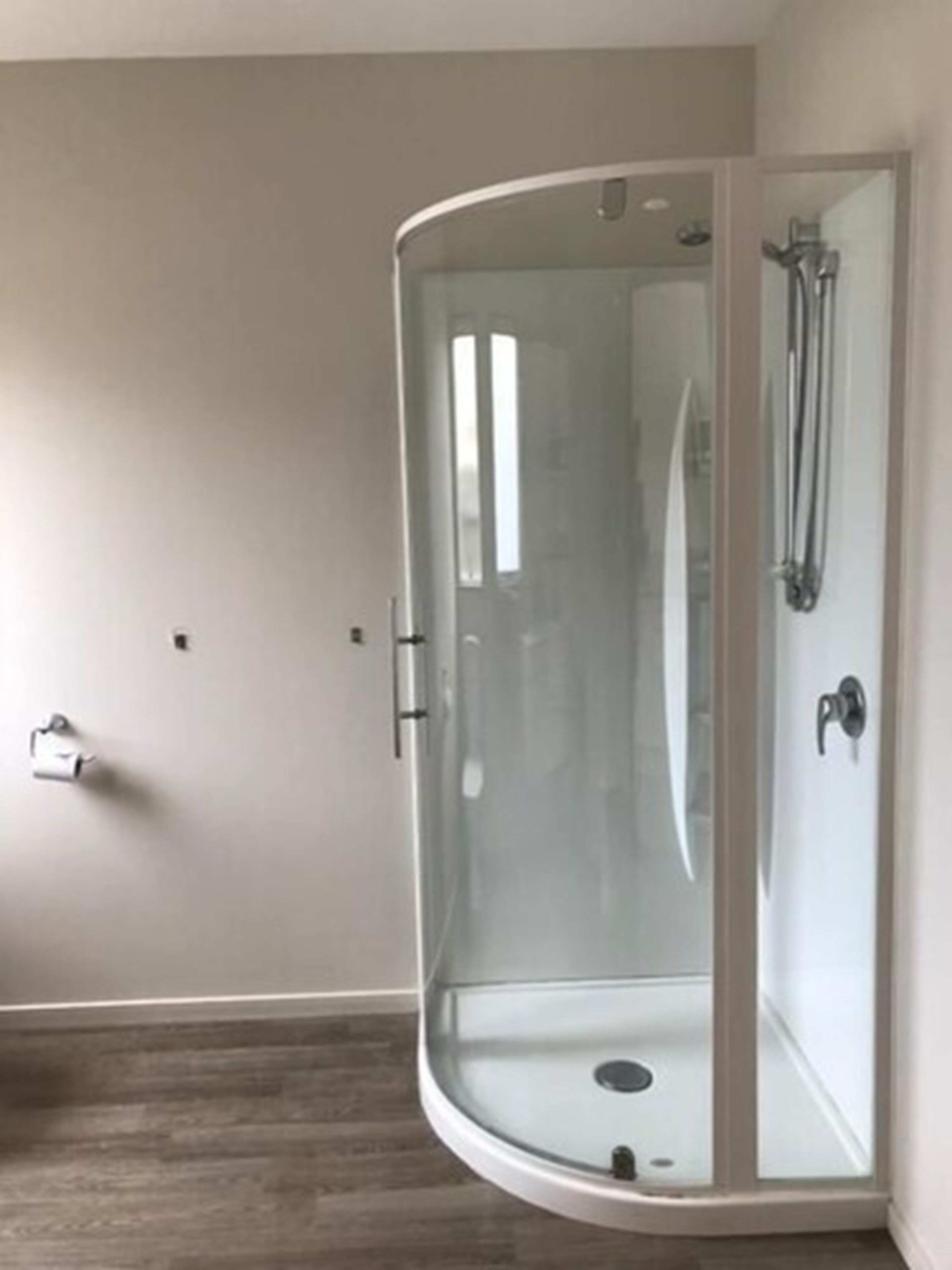 Ducha con cerramiento de vidrio y estructura blanca, suelo de madera y pared lisa.
