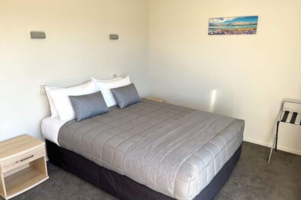 A single bed with a grey quilt, a wooden nightstand, and a small bedside table.