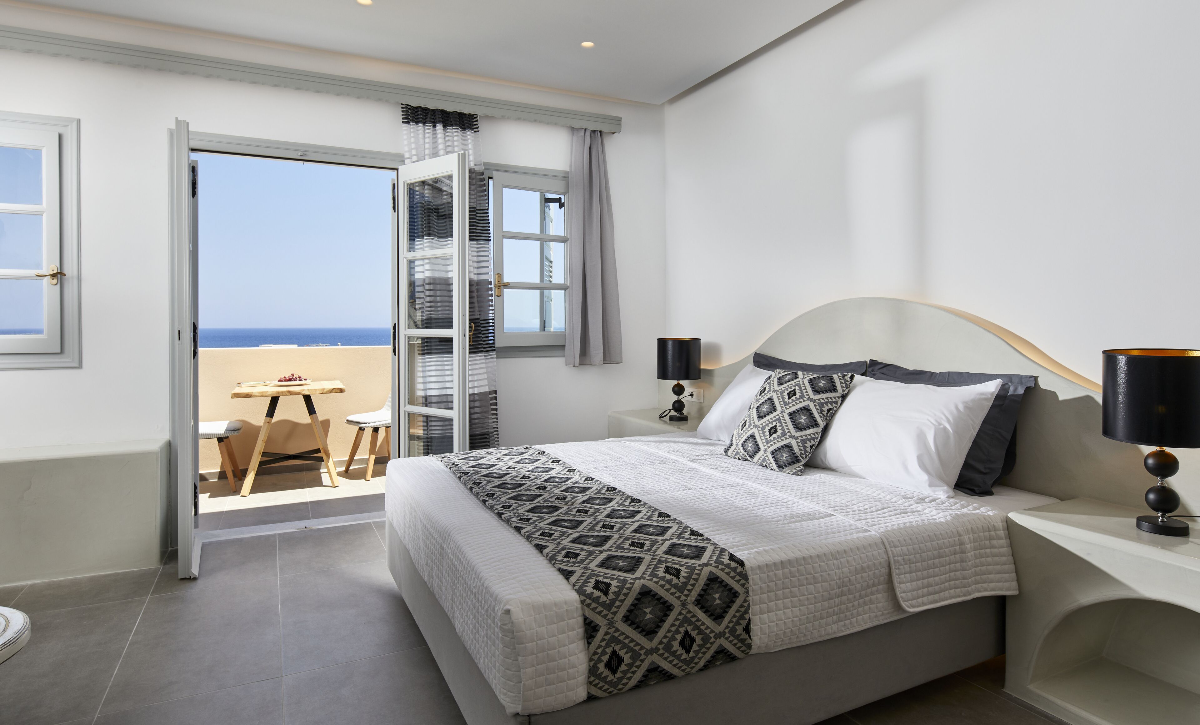 A bedroom with a large bed, two bedside lamps, a view of the sea through a sliding glass door, and a small table with a chair outside.