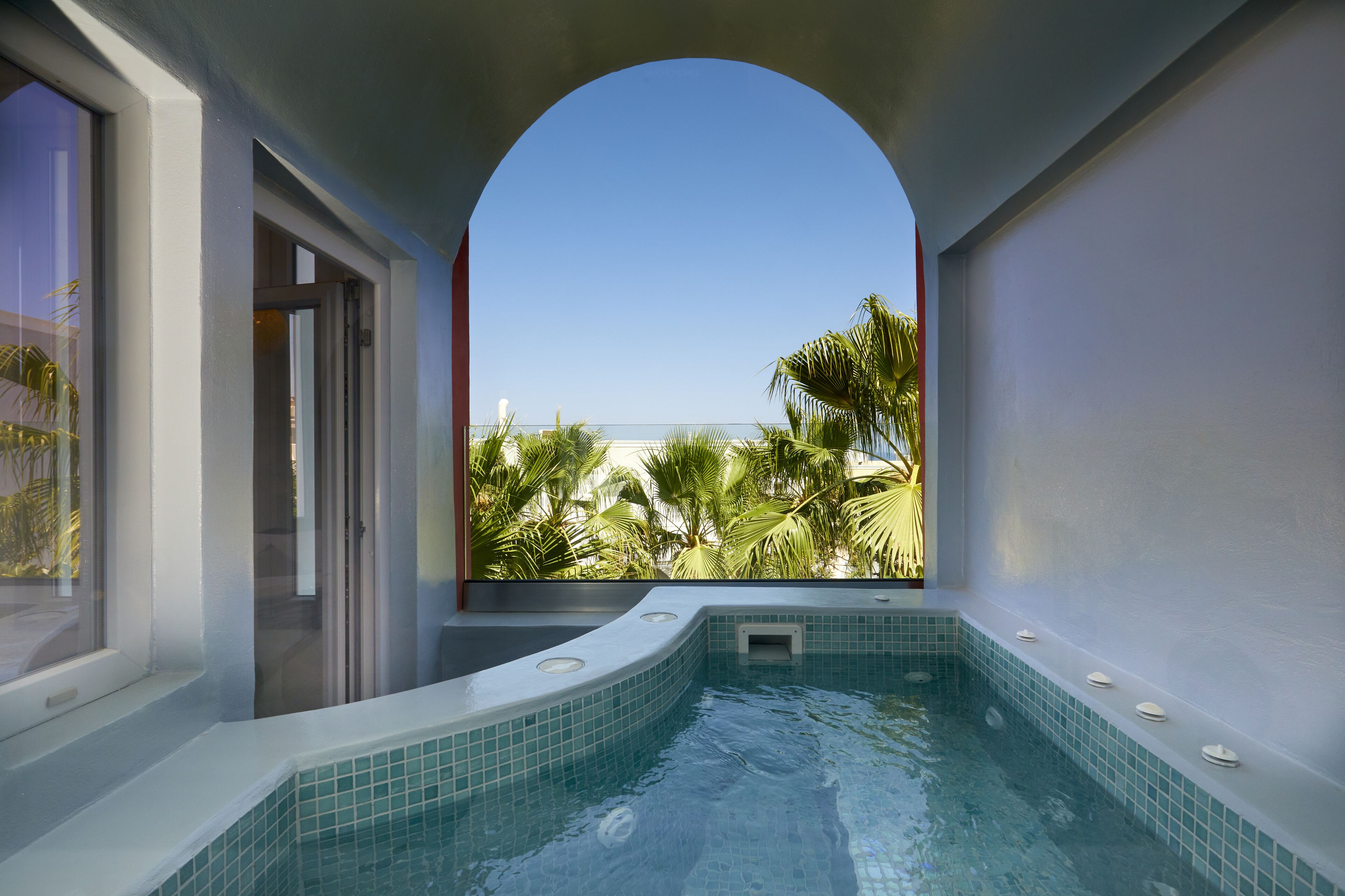 A modern indoor swimming pool with a curved design, a clear view of the ocean, and a tropical garden outside.