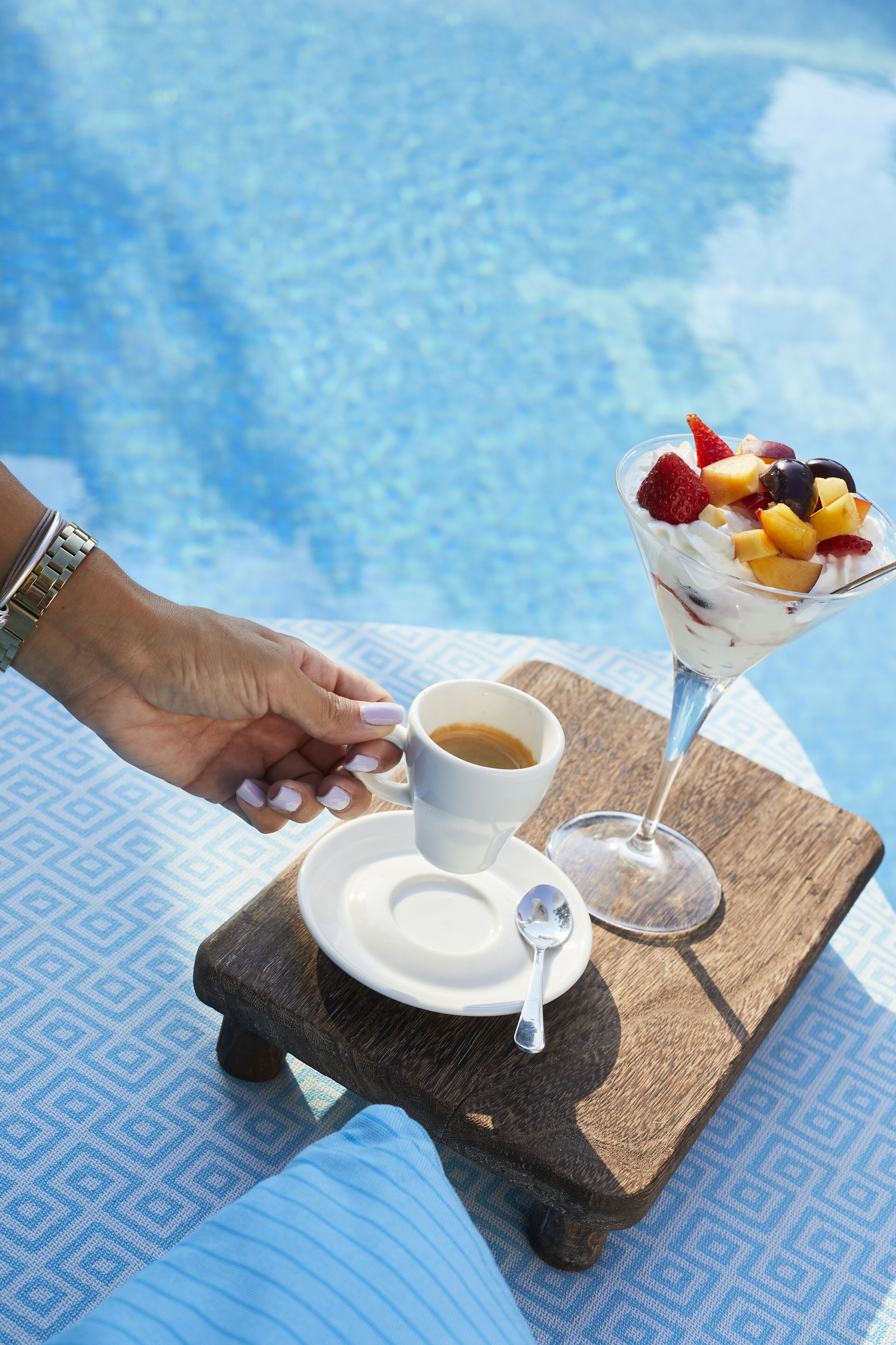 Eine Hand hält eine Kaffeetasse neben einem Desserttablett mit Früchten und einem Glas Saft, auf einem Holztisch am Pool.