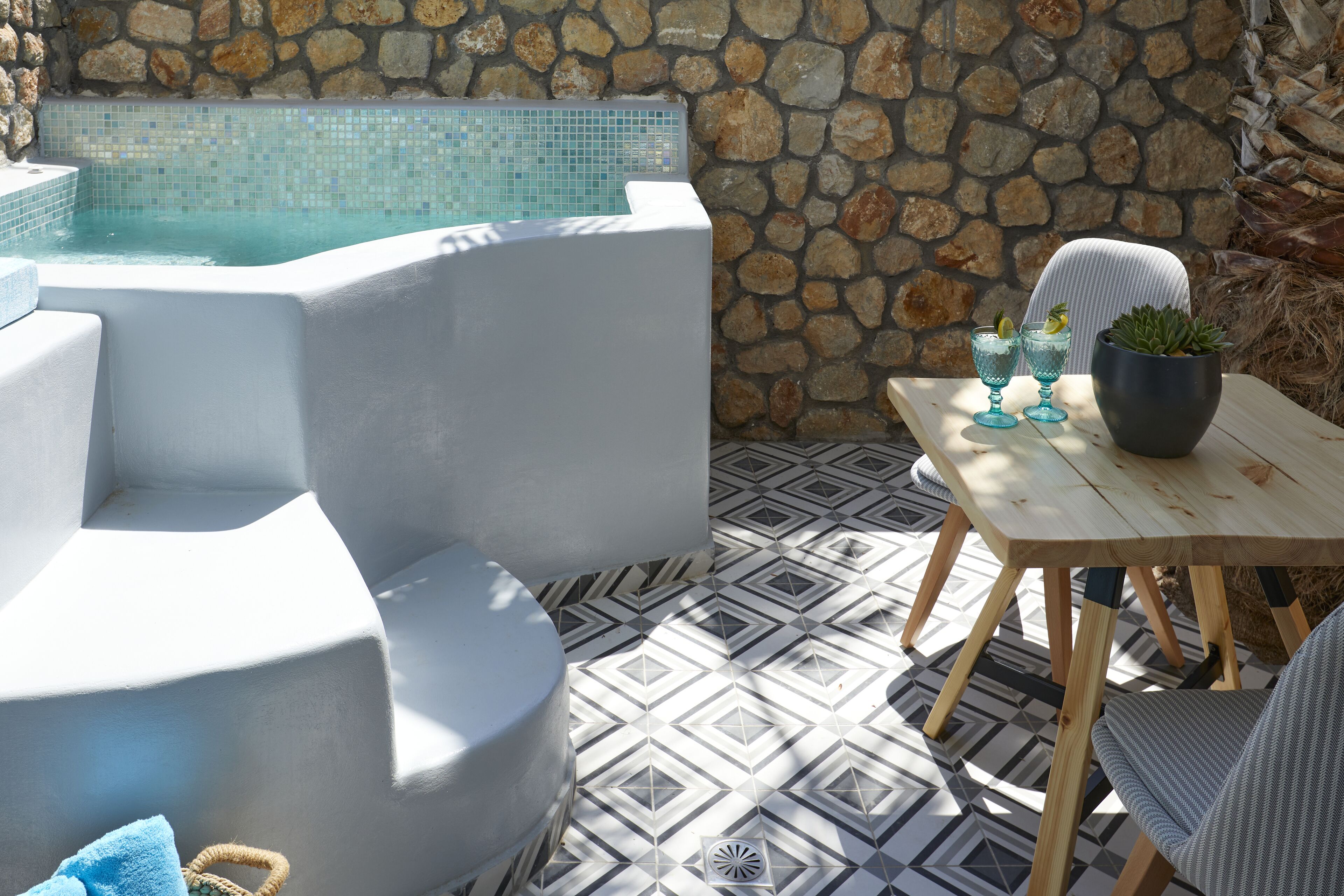Un muro de piedra, una piscina, una silla blanca, una mesa y una planta en maceta.