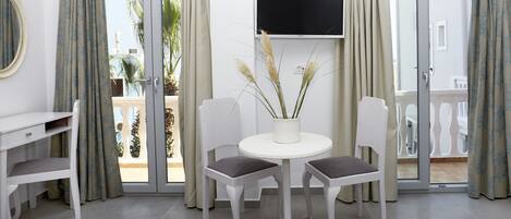 A room with a white desk, two chairs, a small table with a potted plant, a flat-screen TV mounted on the wall, and a sliding glass door leading to an outdoor area.