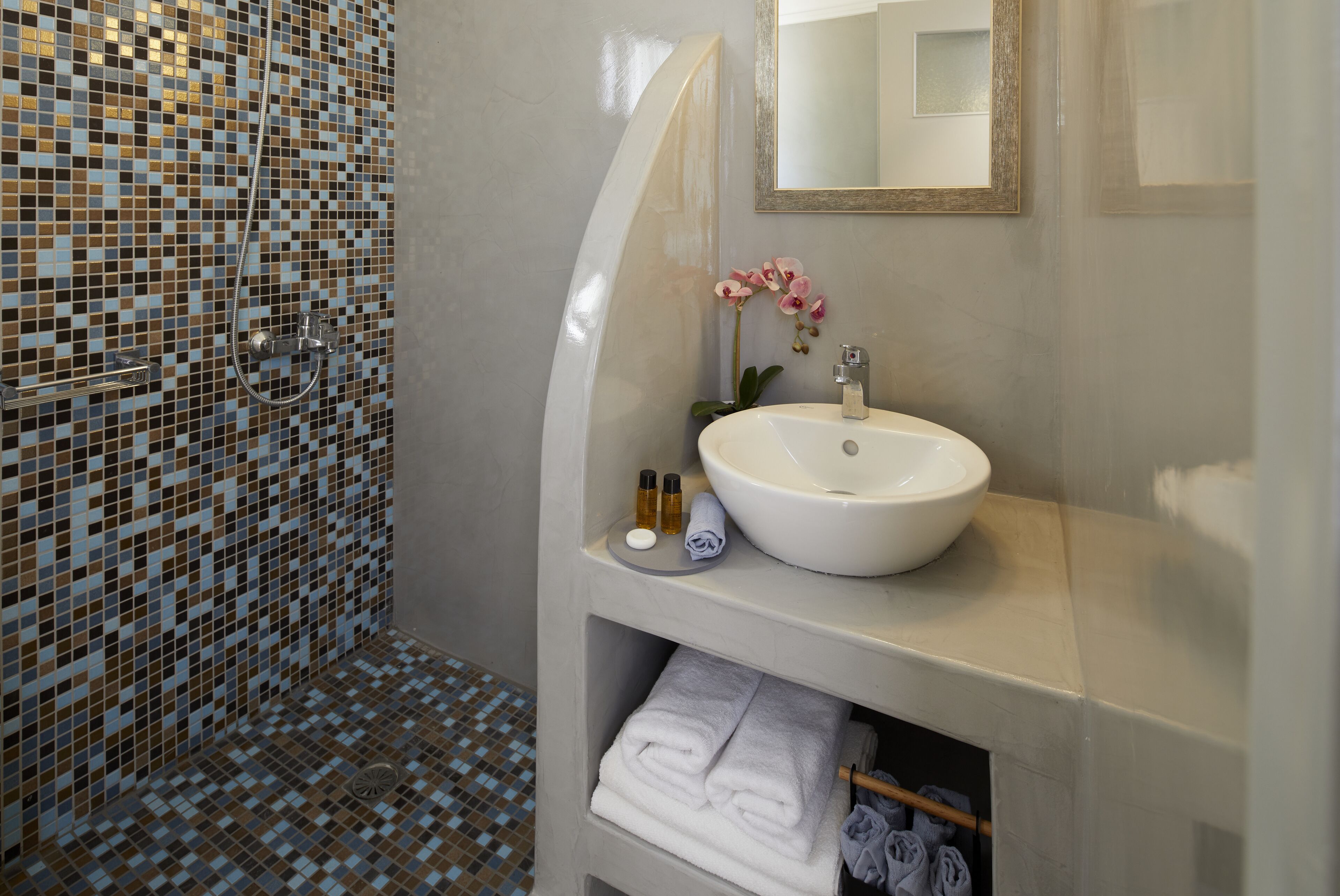 A bathroom with a mosaic tile shower wall, a white sink with a mirror above it, and neatly folded towels on a shelf.