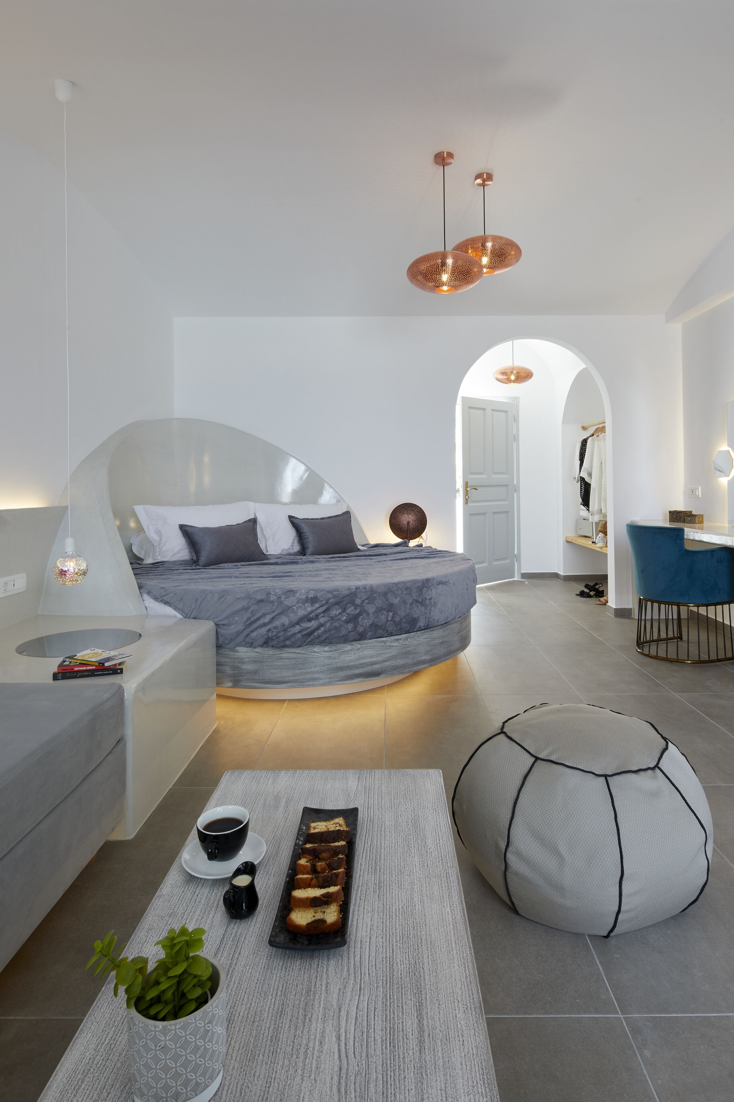 A modern bedroom with a curved bed, a coffee table with a tray of bread, a potted plant, and a seating area with a blue chair.