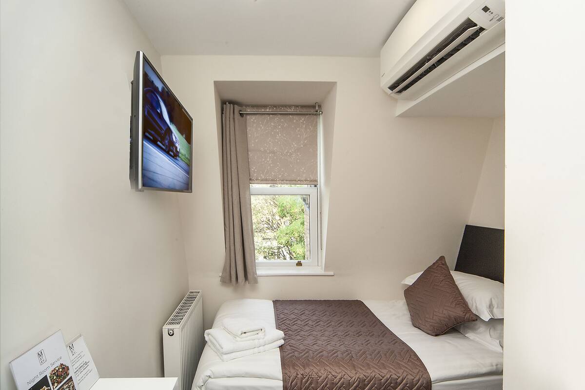 A small, neatly arranged bedroom with a bed, a window with curtains, an air conditioning unit, and a mounted television.