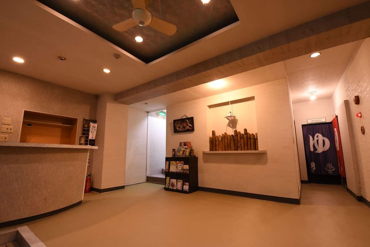 Een hotel lobby met een receptiebalie, een plafondventilator, inbouwverlichting, een wandplank met boeken en een deur met een logo.