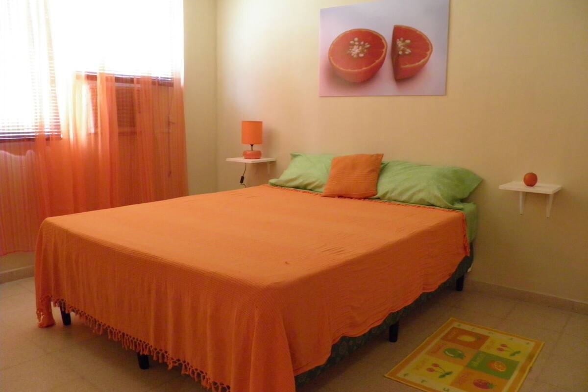 A bedroom with a bed, a nightstand, a lamp, a wall art of fruit, and a fruit-themed rug.