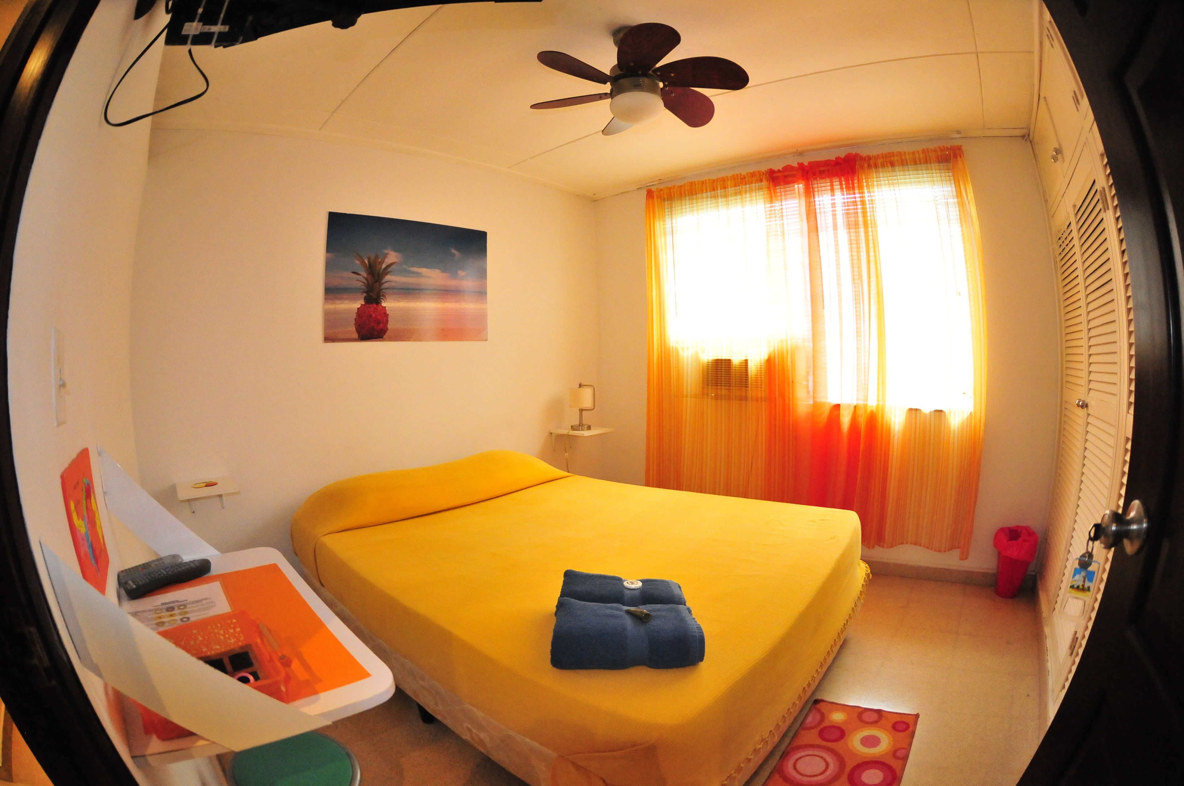 A room with a bed, a ceiling fan, a window with curtains, a painting on the wall, and a small table with items on it.