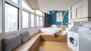 Habitación de hotel compacta con cama, zona de estar, lavadora y ventanas amplias con vista a la ciudad.