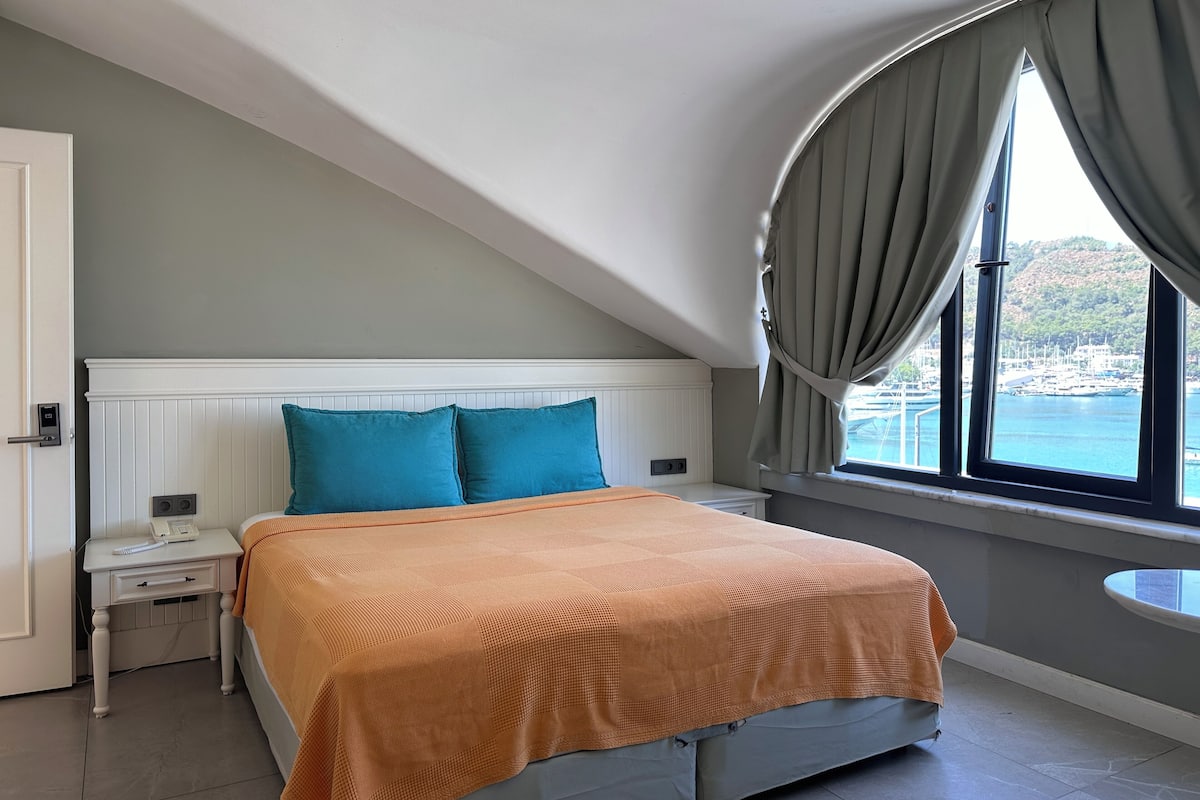 A bedroom with a bed, a nightstand, a telephone, a window with curtains, and a view of a marina.