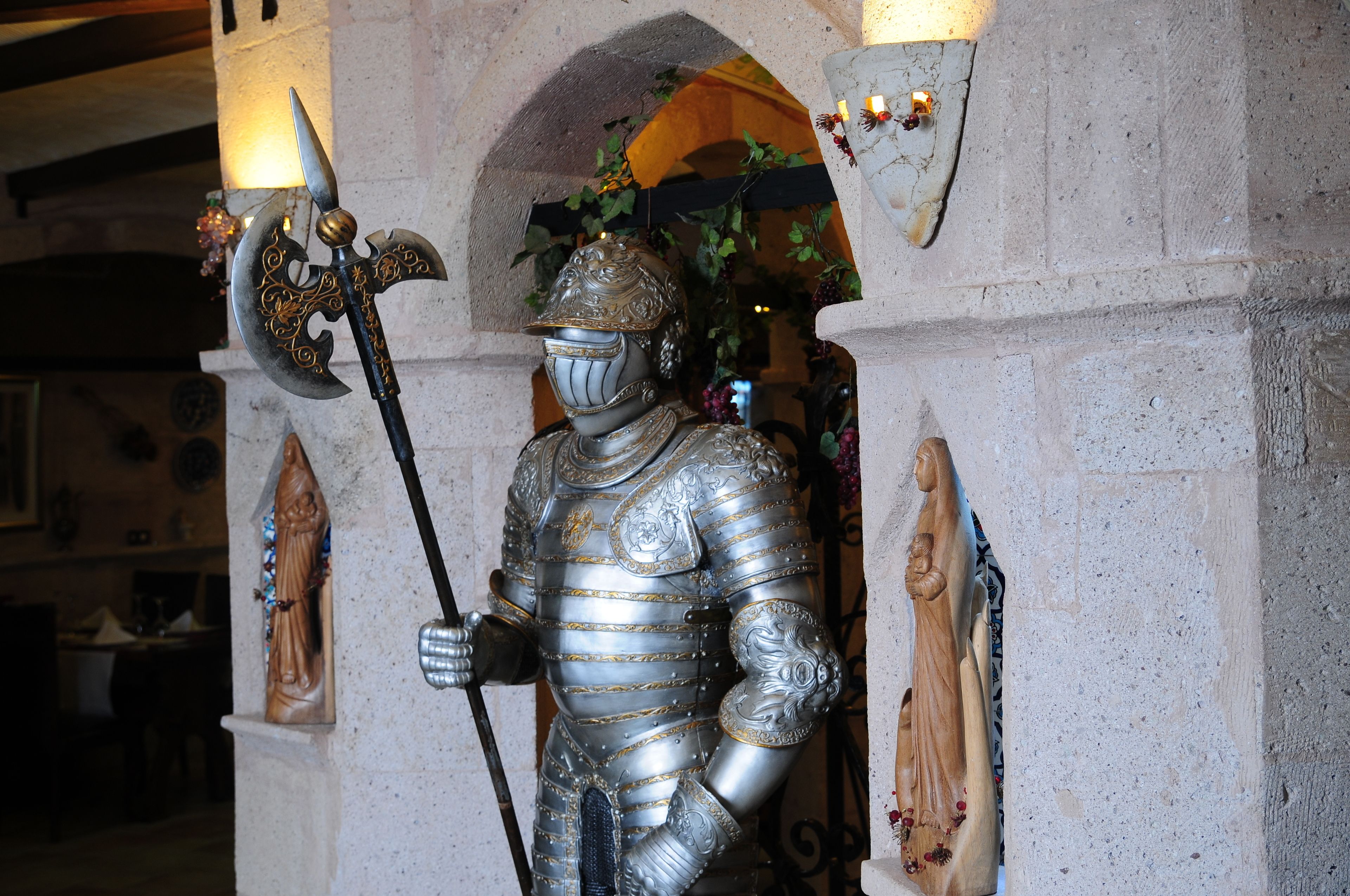 A knight statue in armor holding a spear, set against a stone wall with a decorative arch and a small statue to the left.