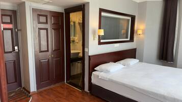 Una camera d'albergo con pavimento in legno, un letto con biancheria da letto bianca, una testiera in legno, uno specchio grande e un bagno visibile attraverso una porta aperta.