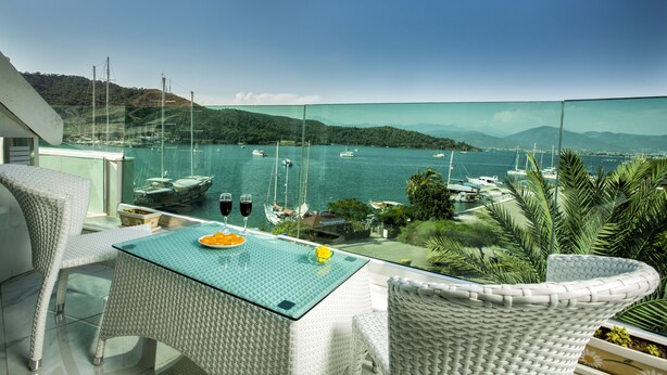 A balcony with a glass table and wicker chairs overlooking a marina with boats. - Ata Park Hotel (Fethiye)
