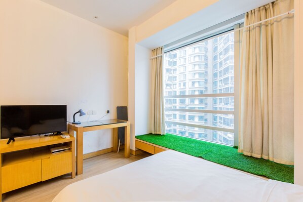 A hotel room with a bed, a TV on a wooden stand, a desk, and a window with a view of a building.
