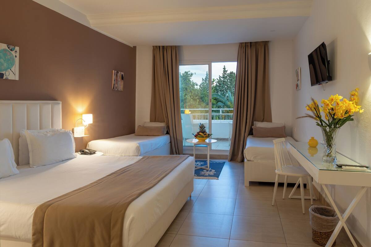 A hotel room with a bed, a desk, a chair, a small table with fruit, and a balcony with curtains.