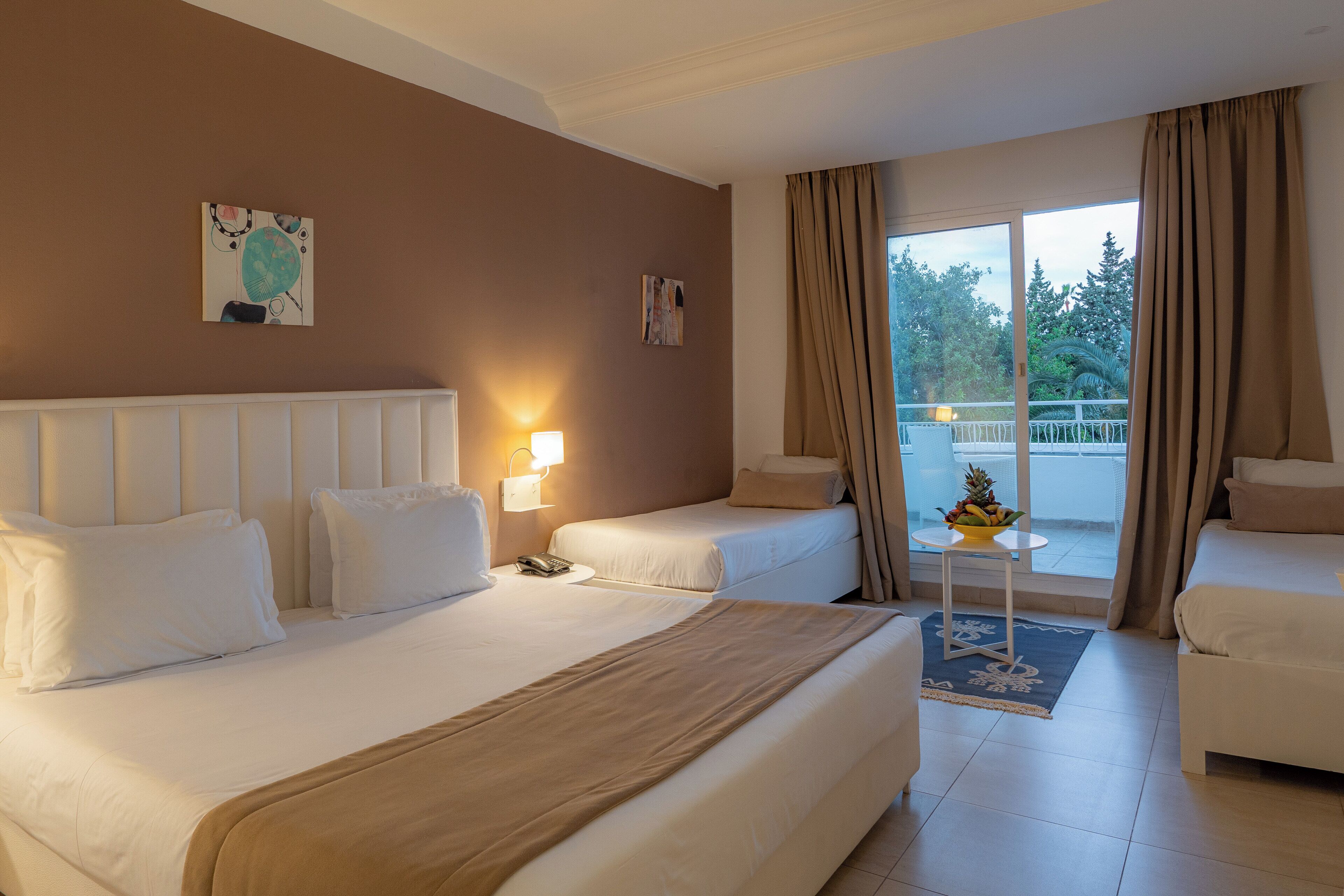 A hotel room with a large bed, two smaller beds, a balcony with curtains, and a small table with a fruit bowl.