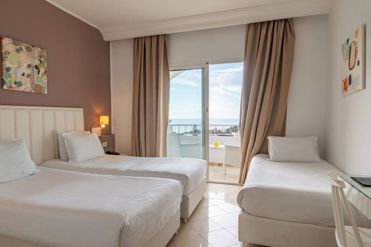 A hotel room with two beds, a balcony with a view, and a small table with a lamp.