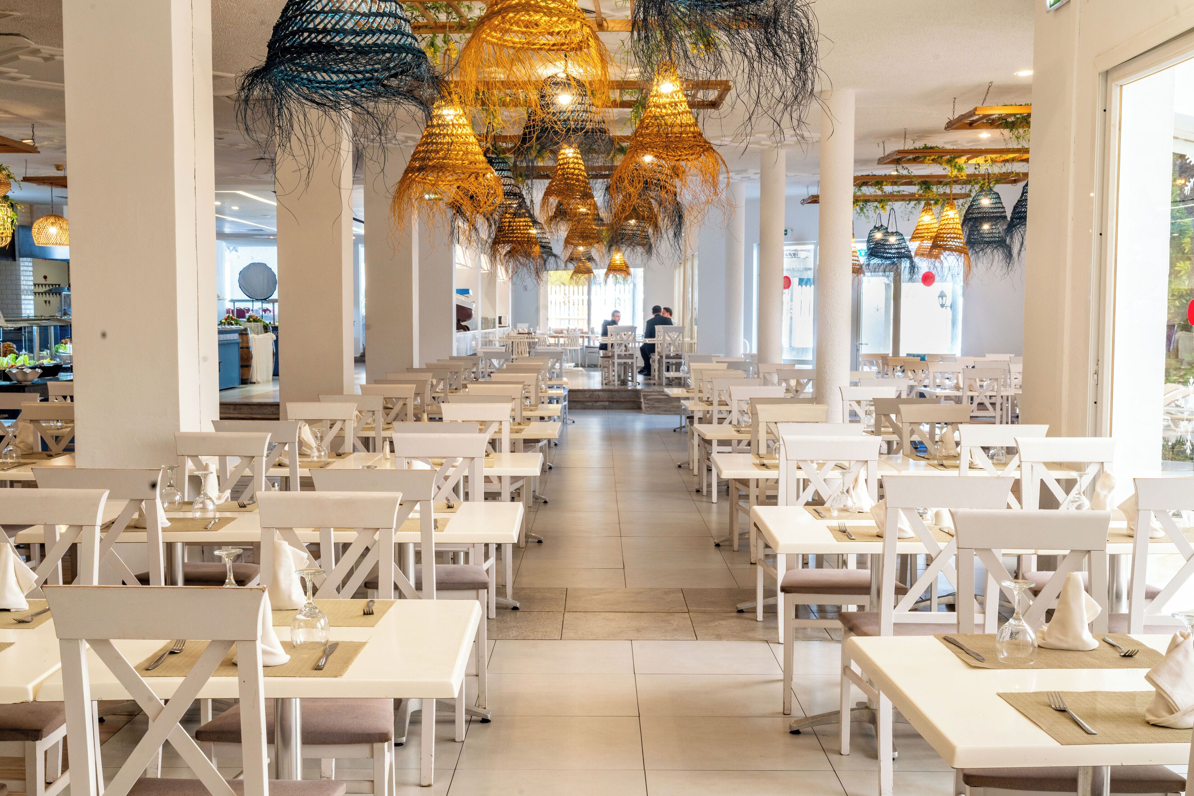 A spacious restaurant with white tables and chairs, a central aisle, and hanging decorative lights.