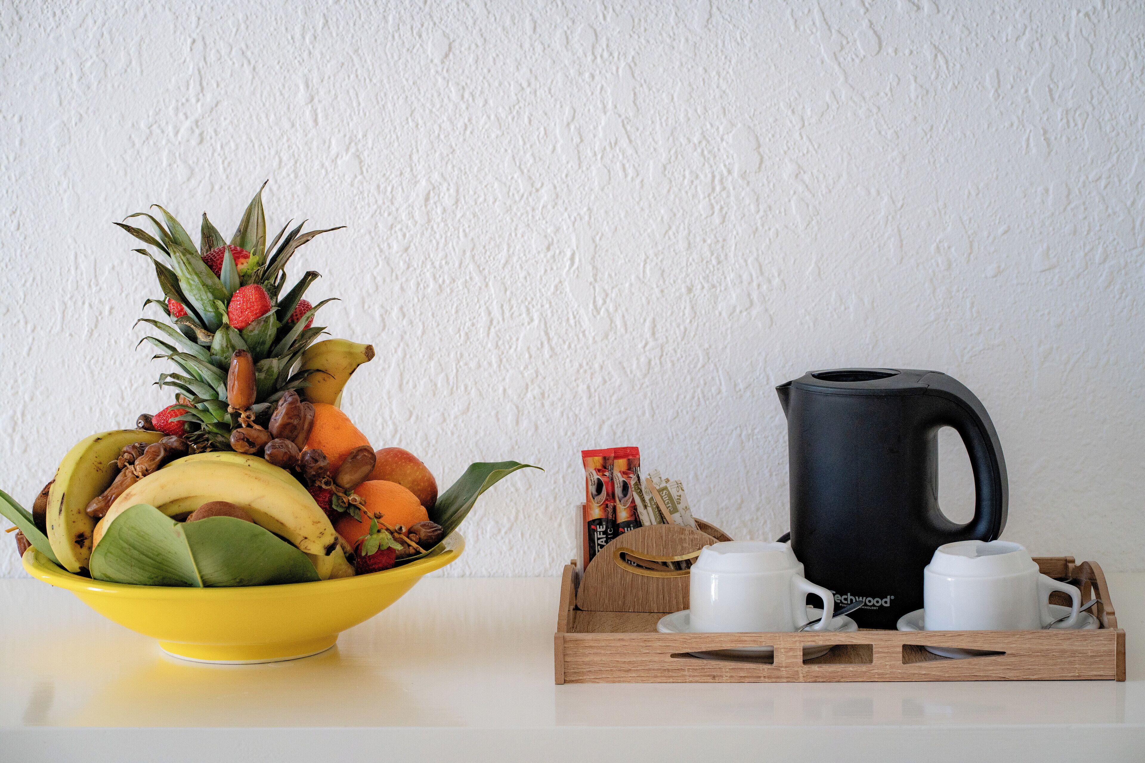 Un tazón amarillo con frutas y una cafetera negra sobre una bandeja con tazas.