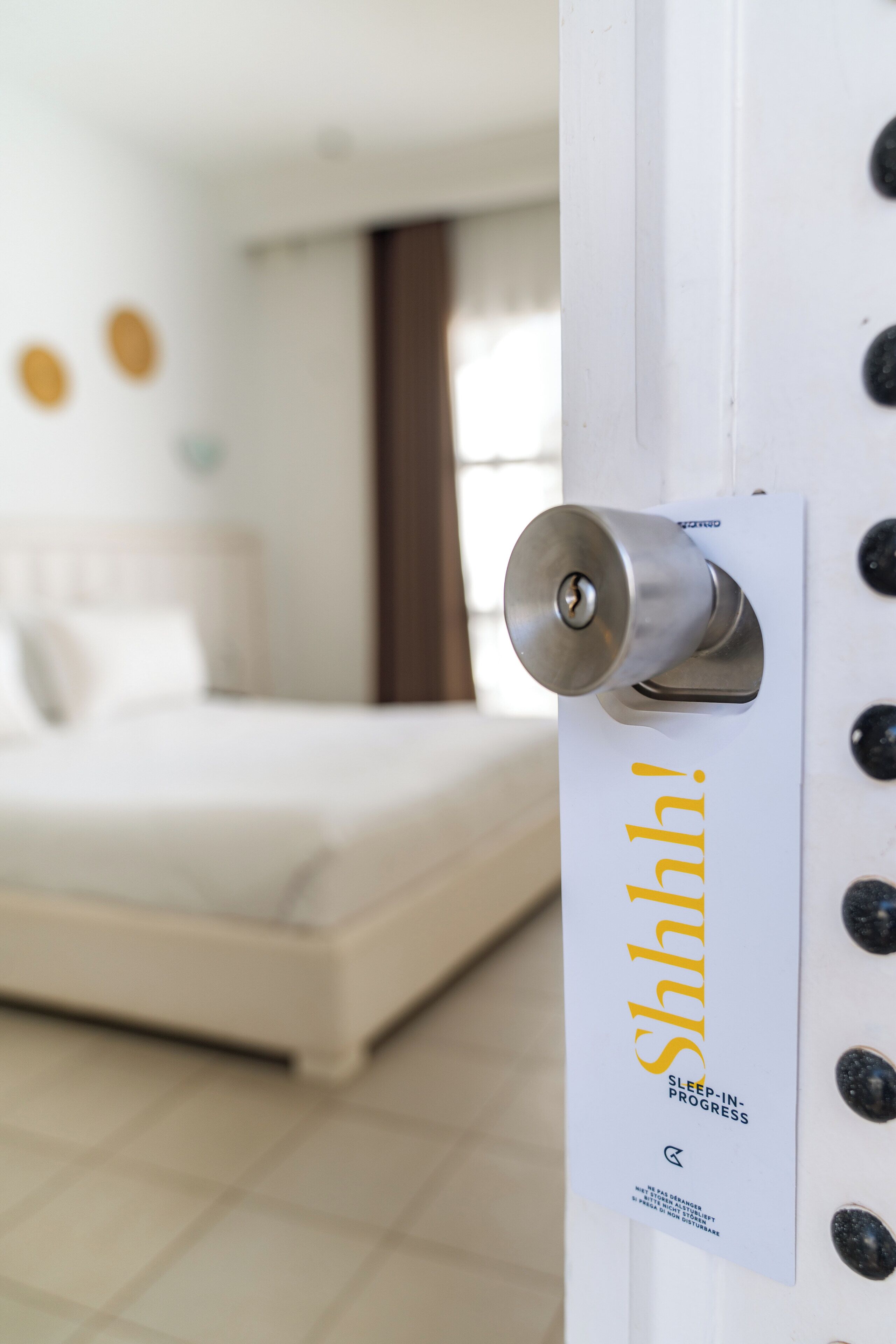 A hotel room with a bed, a door with a keyhole, and a sign with the text 'Shhh! SLEEP-INPROGRESS'.