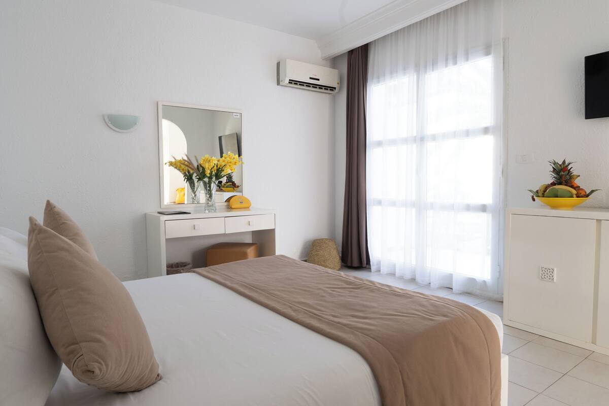 A hotel room with a bed, a desk, a mirror, an air conditioner, a window with curtains, and a fruit bowl on the desk.